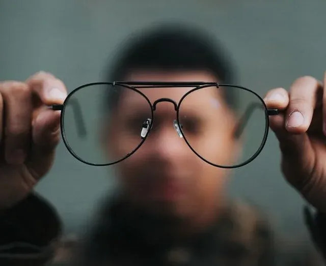 A person holding eyeglasses up to the camera, with their face blurred behind the lenses